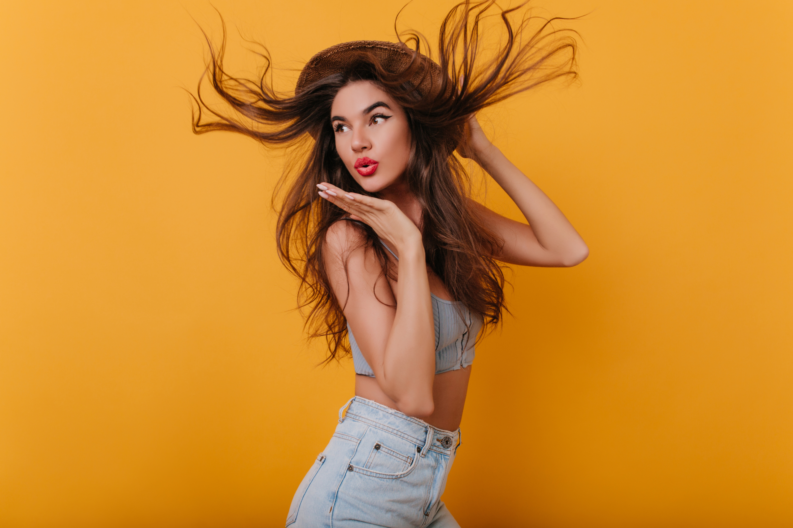 Amazing girl with long straight hair dancing in studio and sending air kiss. Indoor portrait of lovely brunette female model in hat and denim pants isolated on yellow background..